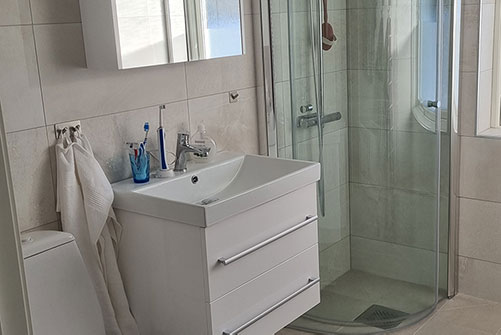 Ett ljust kaklat badrum med vita badrumsmöbler och duschvägg i bakgrunden