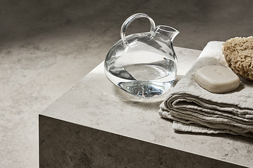 En avlastningsyta i beige sten i ett badrum med handduk, tvål och en vattenkanna i glas ovanpå