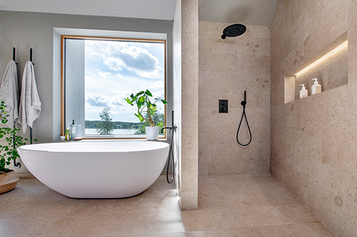 Bad rum med beige sten, svarta detaljer och ett badkar precis vid ett fönster med utsikt över vatten